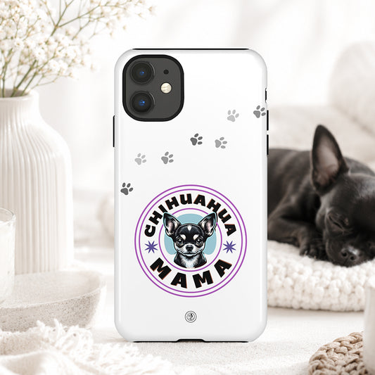 A white phone case featuring a 'Chihuahua Mama' graphic with a chihuahua illustration and paw prints, displayed in a cozy home setting with a sleeping black chihuahua in the background.