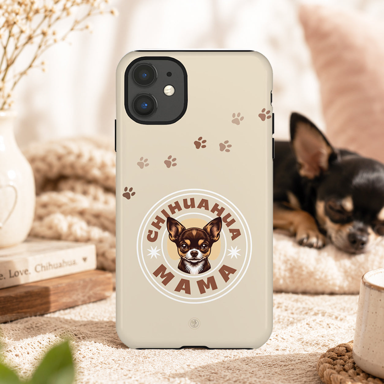 A beige phone case featuring a Chihuahua Mama graphic and paw prints, shown on a dog bed with a small Chihuahua in a cozy home setting.